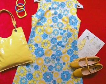 Puede incluir: Un vestido sin mangas con un patrón repetitivo de flores amarillas y azules sobre fondo blanco, acompañado de un bolso de charol amarillo, tres brazaletes de plástico, un par de zapatos Mary Jane amarillos y un patrón de costura de papel.