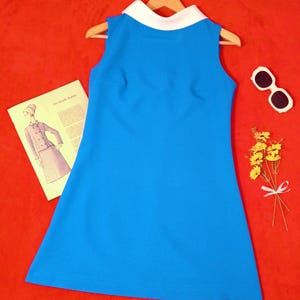 Pode incluir: Um vestido sem mangas azul brilhante com gola branca, apresentado num cabide de madeira. O vestido é acompanhado por um folheto de moldes de costura vintage, óculos de sol com armação branca e um pequeno ramo de flores amarelas.