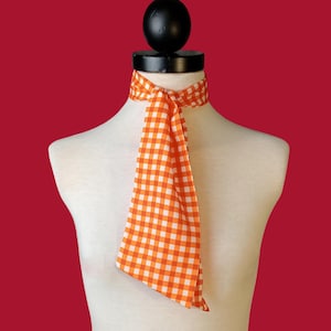 May include: A narrow neck scarf featuring a vibrant orange and white gingham check pattern. The fabric is tied around the neck of a mannequin, with two long, pointed ends hanging down the center of the chest in a simple, straight knot style.