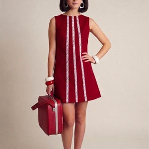 Può includere: Un miniabito rosso senza maniche con finiture in pizzo bianco, abbinato a scarpe rosse e una valigia vintage. L'abito presenta una classica silhouette a trapezio, che ricorda lo stile retrò degli anni '60. La valigia ha bordi argentati.