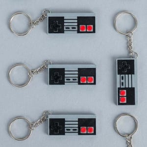 May include: A collection of gray and black keychains shaped like retro video game controllers. Each keychain has a silver ring and chain. The controllers feature a directional pad, buttons, and red accents, reminiscent of classic gaming consoles.