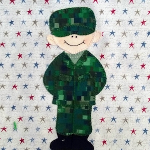 May include: A felt appliqué of a smiling soldier wearing a green camouflage uniform and a black hat. The soldier is standing on a white background with red and blue stars.