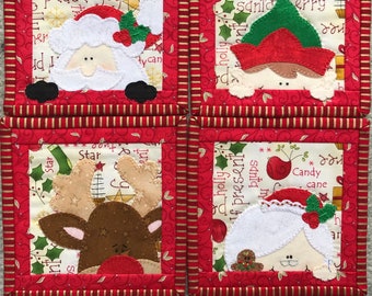 May include: Four red and gold Christmas-themed coasters with appliqued characters. The coasters feature Santa, an elf, a reindeer, and a snowman. The coasters are decorated with holly, stars, and the words "Happy Holidays", "Peace", "Joy", "Star", "Elf Present", "Holly", "Candy Cane", "Santa", "Tree", "Merry", and "Green".