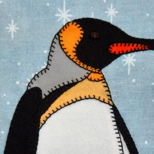 Puede incluir: Una aplicación de pingüino de fieltro con detalles en negro, blanco, naranja y gris. El pingüino está de pie sobre un fondo azul con copos de nieve blancos.