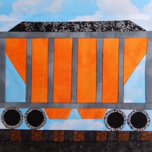 May include: A colorful fabric art piece depicting a train car. The car features orange panels, gray supports, and black wheels. The background is a blue sky with white clouds, and the tracks are brown.