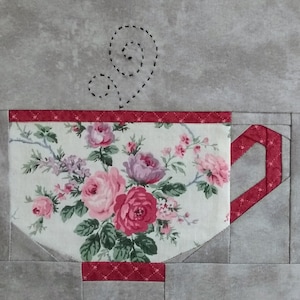 Op de afbeelding: Een rood en wit theekopje met een bloemenpatroon en een rode handgreep. Het theekopje staat op een grijze achtergrond met een zwart gestikt stoomontwerp erboven.