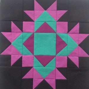 May include: A quilt block with a starburst pattern in pink, teal, and burgundy. The block is set against a black background.