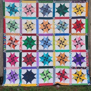 May include: A handmade quilt featuring a repeating star pattern in various colors, including blue, green, pink, and yellow. The stars are set against a white background, with each star block framed by a contrasting border. The quilt is likely made of cotton fabric.