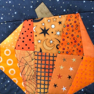 May include: A patchwork pumpkin with various orange fabrics, each with a unique pattern. The patterns include polka dots, stars, moons, and spiderwebs. The background is a dark blue fabric with white stars, and the stem is a tan fabric.