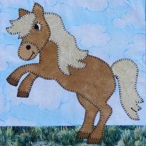 May include: A brown and white felt horse applique with a stitched outline. The horse is standing on its hind legs with its front legs raised in the air. The horse has a long mane and tail. The background is a light blue with white clouds.