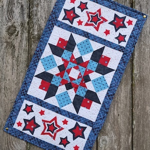 Puede incluir: Un camino de mesa acolchado azul y blanco con un diseño de estrellas rojo, blanco y azul. El camino de mesa está hecho de tela con un patrón de cuadros y presenta una gran estrella en el centro.