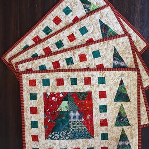 May include: Four Christmas-themed quilted placemats. Each has a cream background with gold snowflakes, red and green squares, and a red striped border. The central design features Christmas trees, adding festive charm.