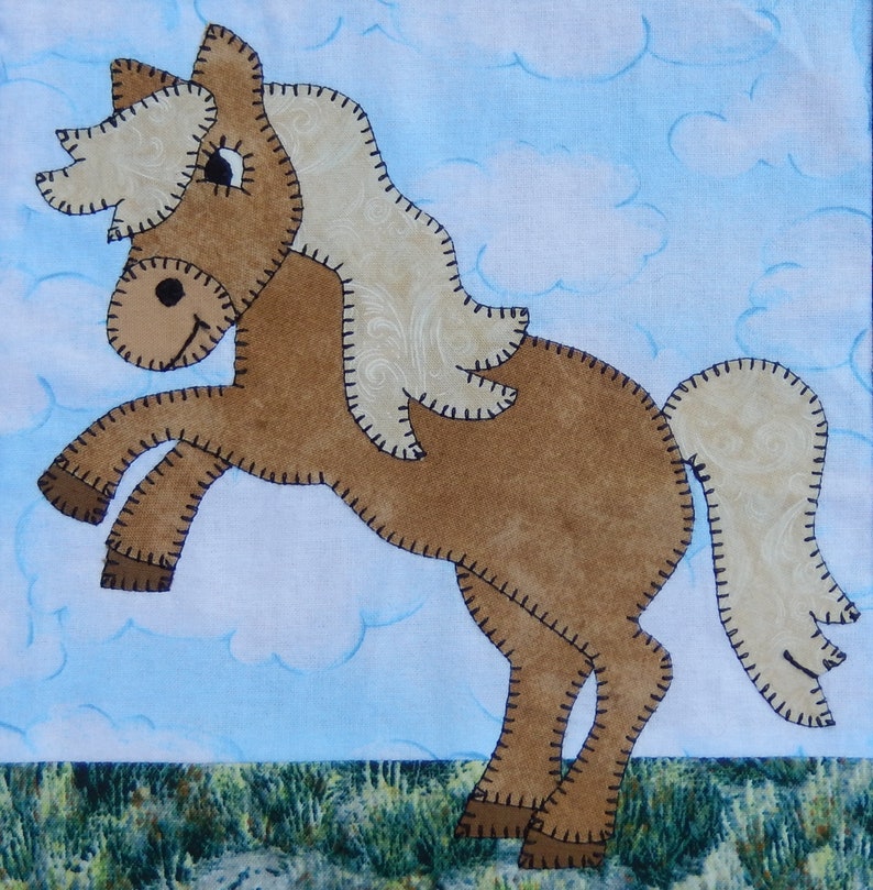 May include: A brown and white felt horse applique with a stitched outline. The horse is standing on its hind legs with its front legs raised in the air. The horse has a long mane and tail. The background is a light blue with white clouds.