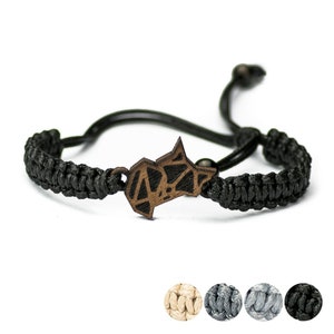Puede incluir: Pulsera trenzada negra con un colgante geométrico de madera. El colgante tiene la forma de un animal estilizado. La pulsera es ajustable y está disponible en varios colores.