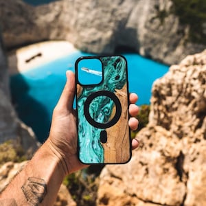 May include: A phone case with a turquoise and brown wood design. The case has a black ring on the back. The phone case is being held in front of a scenic view of a beach and cliffs.