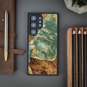 Può includere: Una custodia per telefono nera con un design unico. La custodia presenta una combinazione di legno e resina, con un motivo a vortice verde e oro. La parte inferiore della custodia ha un design a venature del legno. La custodia è accanto a un taccuino in pelle e una penna.