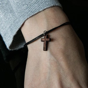 Puede incluir: Un brazalete de cordón negro con un colgante de cruz de madera. La cruz es de color marrón oscuro y tiene un diseño sencillo.