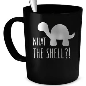 Op de afbeelding: Een zwarte koffiemok met een wit silhouet van een schildpad en de tekst "What the shell?" erop gedrukt.