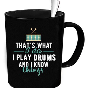 Op de afbeelding: Zwarte koffiemok met een witte handgreep. De mok heeft een afbeelding van een drumstel met drumsticks en de tekst "That's what I do - I play drums and I know things".