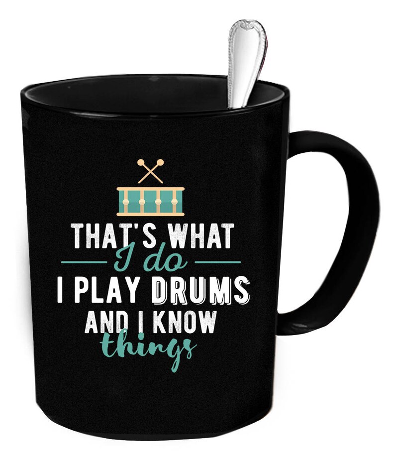Op de afbeelding: Zwarte koffiemok met een witte handgreep. De mok heeft een afbeelding van een drumstel met drumsticks en de tekst "That's what I do - I play drums and I know things".