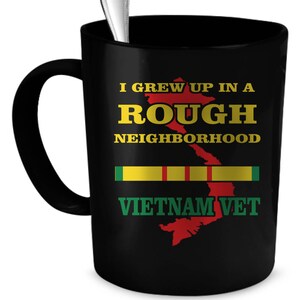 May include: Black coffee mug with a white handle. The mug has a graphic of a red outline of Vietnam with yellow and green stripes and the text "I grew up in a rough neighborhood Vietnam Vet".