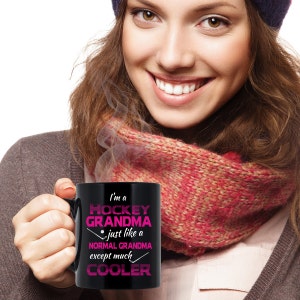 Puede incluir: Taza de caf&eacute; negra con texto blanco que dice "I'm a Hockey Grandma just like a normal grandma except much cooler" en texto rosa. La taza la sostiene una persona que lleva una bufanda roja y blanca.