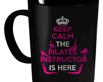 May include: Black ceramic coffee mug with a white crown and the text "Keep Calm The Pilates Instructor Is Here" in pink and white. The mug features a silhouette of a person doing a pilates pose.
