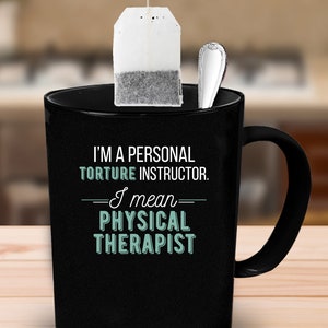 May include: Black coffee mug with a white tea bag hanging from the rim. The mug has the text "I'm a personal torture instructor. I mean physical therapist" printed on it.