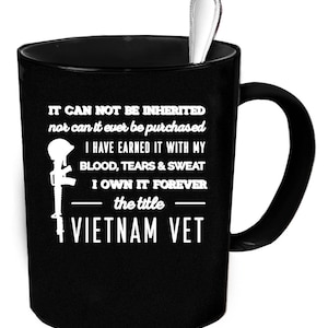 May include: Black ceramic mug with a silver spoon inside. The mug features white text and a graphic of a helmet and rifle, with the words "VIETNAM VET". The text reads "IT CAN NOT BE INHERITED...I OWN IT FOREVER".