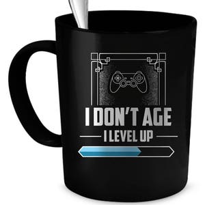 Op de afbeelding: Zwarte koffiemok met een wit ontwerp van een videogamecontroller en de tekst "I don't age I level up" met een blauwe voortgangsbalk.