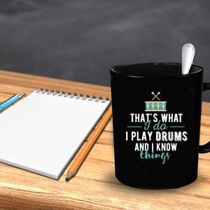 Op de afbeelding: Zwarte koffiemok met een wit ontwerp met de tekst "That's what I do I play drums and I know things". Het ontwerp bevat ook een gestileerde afbeelding van een drumstel met drumsticks.