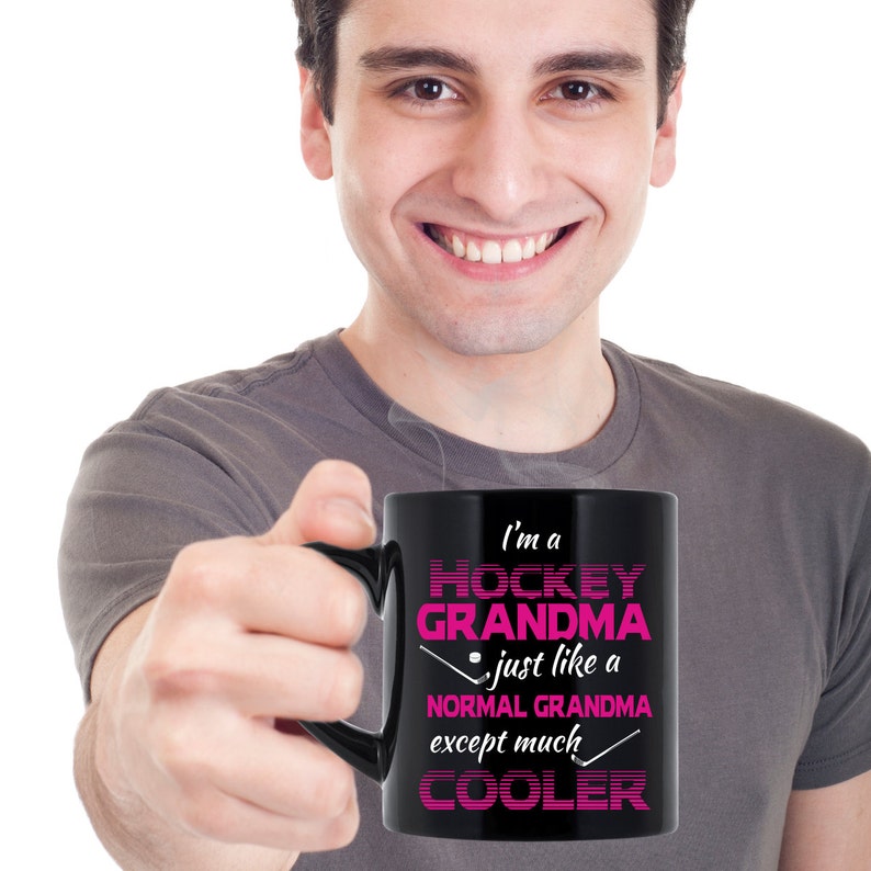Puede incluir: Una taza de caf&eacute; negra con un dise&ntilde;o blanco que dice "Soy una abuela de hockey - como una abuela normal, excepto mucho m&aacute;s genial". El texto est&aacute; en rosa y negro. La taza est&aacute; siendo sostenida por una persona.