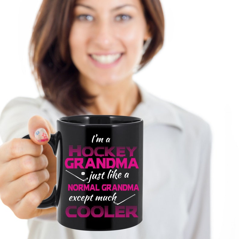 Puede incluir: Taza de caf&eacute; negra con un dise&ntilde;o rosa que dice "Soy una abuela de hockey - como una abuela normal, excepto mucho m&aacute;s genial".