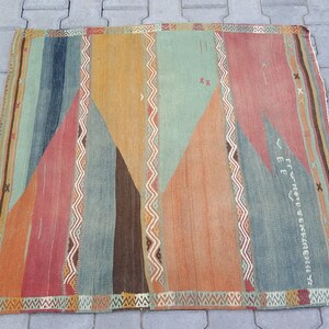 May include: A vintage rug with a geometric pattern in shades of orange, red, blue, and green. The rug has a fringe border and features a faded inscription in the center.