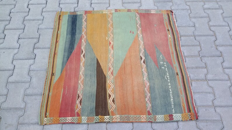 May include: A vintage rug with a geometric pattern in shades of orange, red, blue, and green. The rug has a fringe border and features a faded inscription in the center.