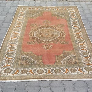 Puede incluir: Una alfombra rectangular de estilo antiguo con un campo central coral y bordes florales ornamentados en beige, marr&oacute;n y naranja. Los intrincados patrones y los bordes con flecos de la alfombra sugieren una est&eacute;tica tradicional y artesanal.