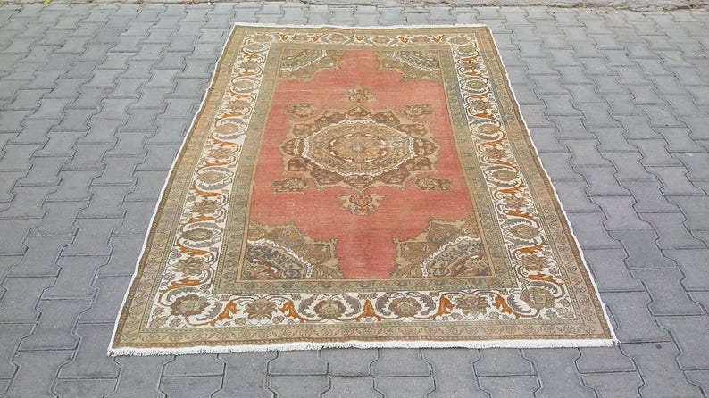Puede incluir: Una alfombra rectangular de estilo antiguo con un campo central coral y bordes florales ornamentados en beige, marr&oacute;n y naranja. Los intrincados patrones y los bordes con flecos de la alfombra sugieren una est&eacute;tica tradicional y artesanal.