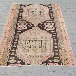 Puede incluir: Una alfombra turca vintage con un patrón geométrico en tonos de rosa, marrón y beige. La alfombra tiene un aspecto descolorido y desgastado, lo que le da un carácter único.