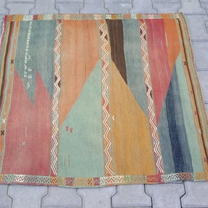 May include: A colorful vintage rug with a geometric pattern in shades of red, orange, green, and blue. The rug has a fringe border and features a repeating pattern of triangles and zigzags.