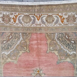 Puede incluir: Primer plano de una alfombra vintage con un campo central rosa y bordes ornamentados. Los bordes presentan dise&ntilde;os florales y geom&eacute;tricos en tonos beige, marr&oacute;n, gris y naranja. La alfombra tiene un borde con flecos y un aspecto desgastado.