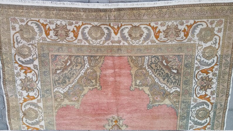 Puede incluir: Primer plano de una alfombra vintage con un campo central rosa y bordes ornamentados. Los bordes presentan dise&ntilde;os florales y geom&eacute;tricos en tonos beige, marr&oacute;n, gris y naranja. La alfombra tiene un borde con flecos y un aspecto desgastado.