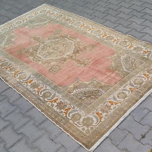 Puede incluir: Alfombra rectangular con un campo central rosa y un borde detallado. La alfombra presenta una paleta de colores beige y marr&oacute;n con dise&ntilde;os florales y geom&eacute;tricos. Se encuentra sobre una superficie de ladrillos grises.