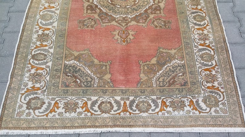 Puede incluir: Una alfombra rectangular de estilo vintage con un campo central de color coral descolorido. La alfombra presenta un borde ornamentado con patrones florales y geom&eacute;tricos en tonos beige, gris y naranja. El dise&ntilde;o de la alfombra es sim&eacute;trico y detallado.