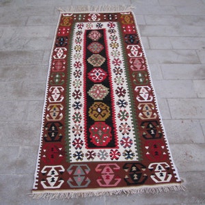 May include: A traditional Turkish rug with a long, rectangular shape. The rug features a colorful geometric pattern in shades of red, green, brown, and white. The rug has a fringe border.