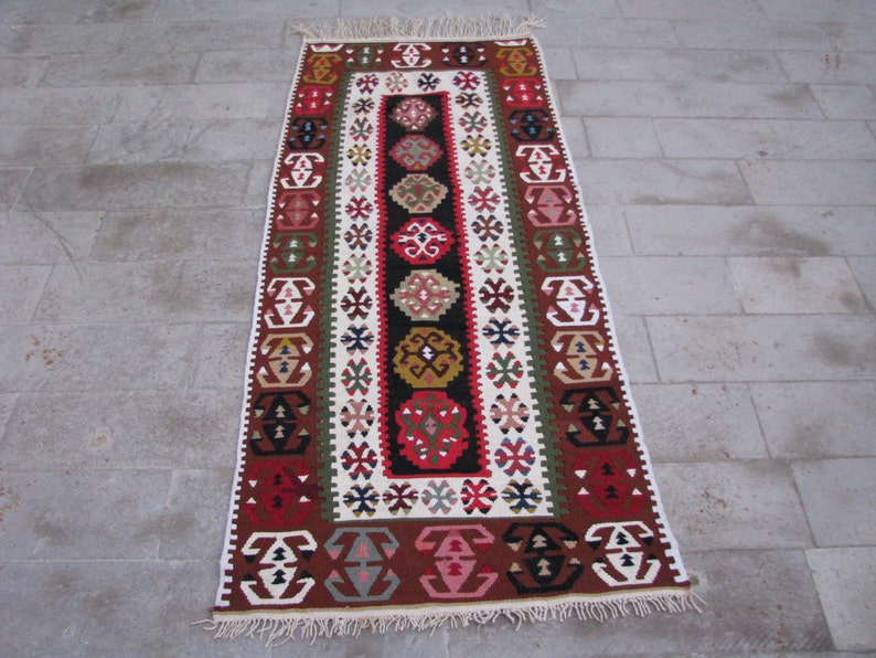 May include: A traditional Turkish rug with a long, rectangular shape. The rug features a colorful geometric pattern in shades of red, green, brown, and white. The rug has a fringe border.