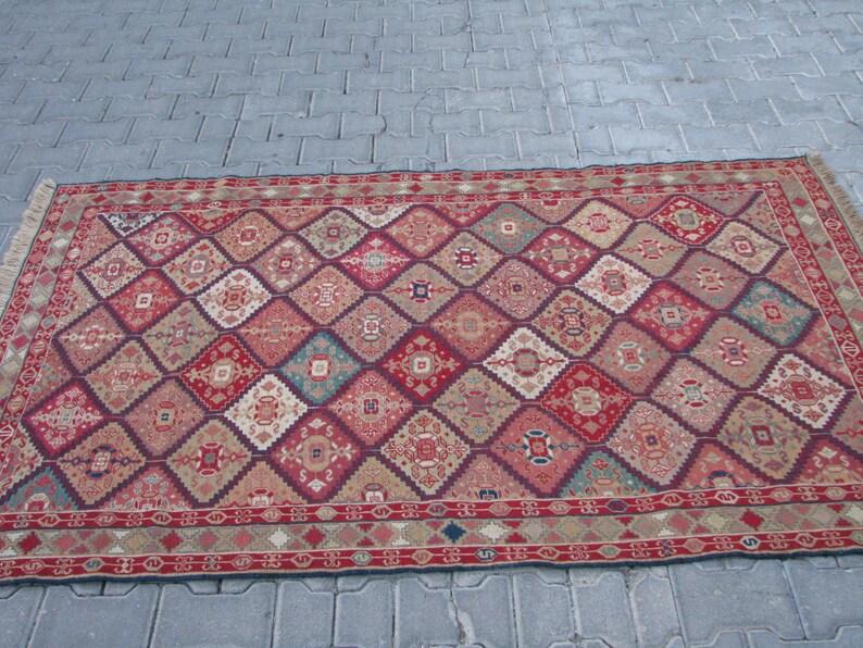 May include: A red, beige, and blue patterned rug with a diamond design. The rug has a fringe border.
