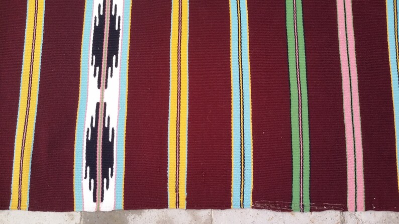 May include: A striped woven rug with a burgundy background. The rug has stripes of yellow, light blue, dark blue, green, and pink.