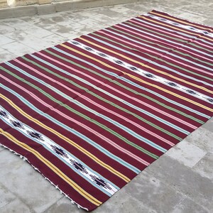 May include: A striped woven rug with a maroon background and yellow, green, white, and blue stripes. The rug has a fringe border.