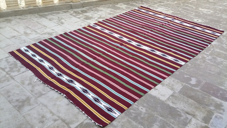 May include: A striped woven rug with a maroon background and yellow, green, white, and blue stripes. The rug has a fringe border.