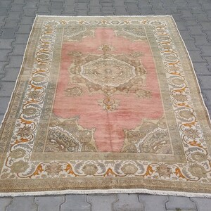 Puede incluir: Una alfombra rectangular vintage con un campo central rosa y dise&ntilde;os de borde intrincados. La alfombra presenta un medall&oacute;n central y detalles en las esquinas en tonos beige, marr&oacute;n y naranja. La alfombra est&aacute; colocada sobre una superficie de ladrillo gris.